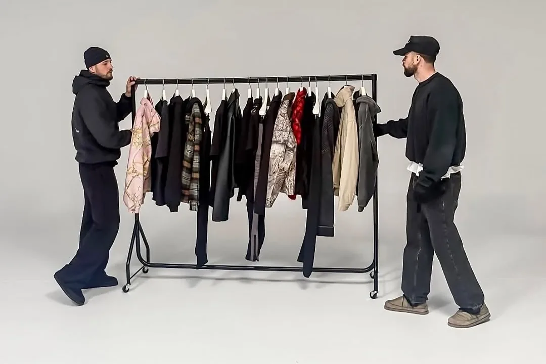 Two people in black wheeling a rail of jackets across a grey studio