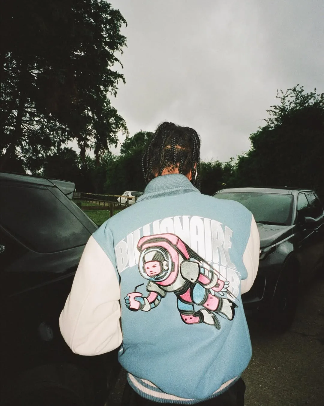A model seen from behind in a pale blue varsity jacket with an astronaut and BILLIONAIRE lettering on the back